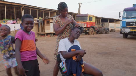 Young-African-woman-braiding-her-hair-outdoors,-with-children-around-her