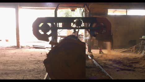 Backlit-silhouette-of-saw-mill-passing-over-large-log-spitting-out-sawdust