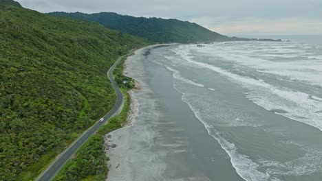 Aerial-view-following-tourist-campervan-traveling-along-the-wild,-rugged-and-remote-West-Coast-with-rolling-waves-on-the-South-Island-of-New-Zealand-Aotearoa