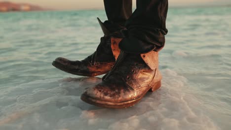 Man-with-old-leather-boots-stands-on-large-salt-outcrop-in-Dead-Sea,-Israel