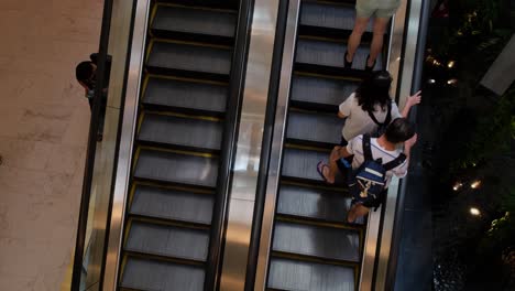 Escalator-at-Emporium-in-Sukhumvit-Road,-revealing-people-going-up-and-one-with-a-backpack,-empty-going-down,-while-a-person-is-seen-pacing-while-looking-at-this-mobile-phone,-Bangkok,-Thailand