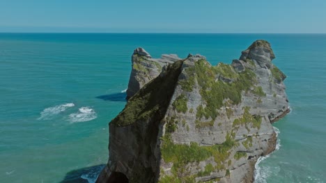 Aerial-flight-over-wild,-rugged-rock-formation-with-arch-in-the-Tasman-Sea-at-popular-tourist-destination-of-Wharariki-Beach-at-Cape-Farewell-in-Nelson,-South-Island-of-New-Zealand-Aotearoa