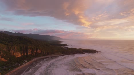 Reverse-aerial-drone-view-of-sunset-overlooking-Pancake-Rocks-on-the-coastline-of-Punakaiki-coastal-landscape,-West-Coast-of-South-Island,-New-Zealand-Aotearoa