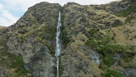 Picturesque-aerial-view-of-rugged,-rocky-and-mountainous-landscape-with-a-free-flowing-narrow-waterfall-in-the-remote-outdoor-wilderness-of-New-Zealand-Aotearoa