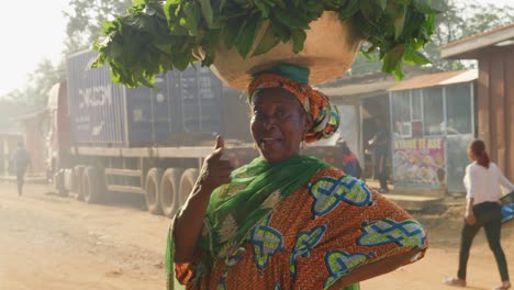 Beautiful-African-woman-in-orange-dress-smiles-balancing-basket-of-greenery-on-head