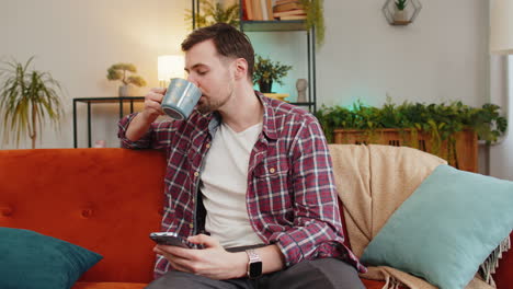 Ein-Glücklicher-Junger-Mann-Nutzt-Sein-Smartphone,-Um-In-Den-Sozialen-Medien-Zu-Chatten,-Während-Er-Zu-Hause-Heißen-Kaffee-Aus-Der-Tasse-Trinkt