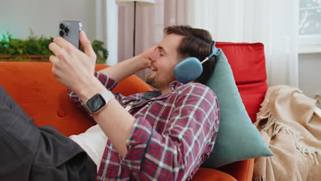 Man-in-wireless-headphones-listening-energetic-dancing-music-on-smartphone-relaxing-lying-on-sofa