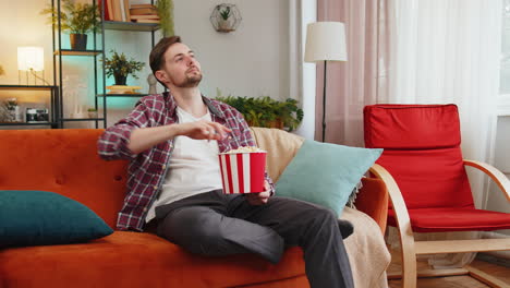 Happy-man-eating-popcorn-watching-interesting-tv-serial-sport-game-online-movie-sits-on-home-sofa