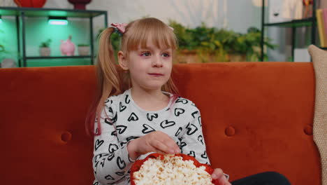 Funny-little-child-girl-watching-comedy-video-film-on-tv,-eating-popcorn-on-comfortable-sofa-at-home