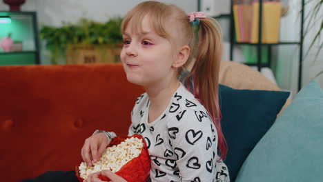 Funny-children-girl-watching-comedy-video-film-on-tv,-eating-popcorn-on-comfortable-sofa-at-home