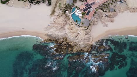 Aerial-birdeye-view-of-a-gorgeous-coastline-with-turquoise-sea-while-the-waves-break-on-the-rocks-right-on-the-beach-with-a-beautiful-villa-with-swimming-pool-on-a-trip-through-mexico-at-chileno-beach