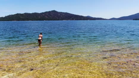 Drone-video-capturing-a-person-swimming-in-a-stunning-blue-lake,-framed-by-a-backdrop-of-lush-green-pine-covered-mountains-in-Whiskeytown-Reservoir,-in-California