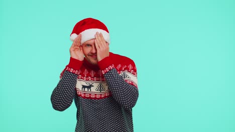 Man-in-sweater-Santa-Christmas-hat-fooling-around-having-closing-eyes-with-hand-and-spying-through