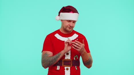Mann-Mit-Weihnachts-T-Shirt-Und-Hut-Tippt-Mit-Seinem-Mobiltelefon-Einen-Neuen-Beitrag-Im-Internet-Ein,-Kauft-Online-Ein-Und-Stöbert