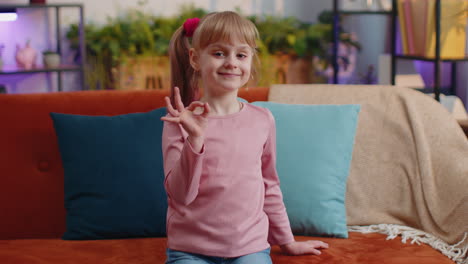 Portrait-of-little-child-girl-kid-alone-on-sofa-at-home-showing-ok-gesture,-like-sign,-positive-good