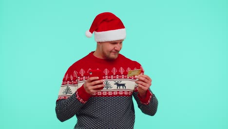 Man-in-Christmas-sweater-using-credit-bank-card,-smartphone-while-transferring-money-shopping-online