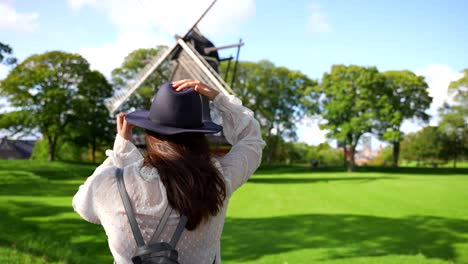 Rückansicht-Einer-Frau,-Die-An-Einem-Windigen-Tag-In-Dänemark-Auf-Die-Windmühle-Blickt-Und-Einen-Hut-Aufsetzt