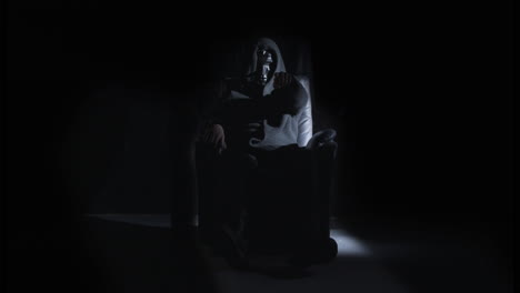 Man-with-scary-mask-sitting-on-chair-in-dark-room-pointing-at-camera