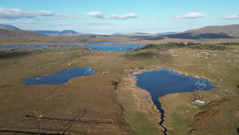 Aerial-view-over-a-landscape-with-several-blue-ponds,-barren-highland-terrain-and-majestic-mountain-ranges-in-background-during-a-travel-through-the-connemara-screebe,-furnace-road-in-galway-ireland
