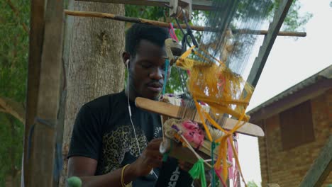 Glücklicher-Afrikanischer-Handwerker,-Der-Kente-Textilien-Auf-Einem-Rustikalen-Webstuhl-Webt-Und-Dabei-Musik-Hört