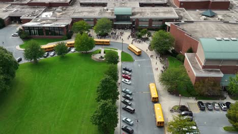Estudiantes-En-Los-Edificios-Del-Campus-De-Una-Escuela-Pública-Mientras-Los-Autobuses-Escolares-Llegan-Para-Transportar-A-Los-Estudiantes