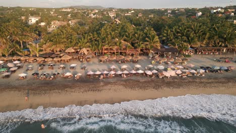 Drone-approaching-la-Punta-in-Zicatela-beach-Puerto-Escondido-Mexico-Oaxaca-coastline-with-people-sunbathing-at-sandy-tropical-beach-with-palm-tree-and-resort-paradise