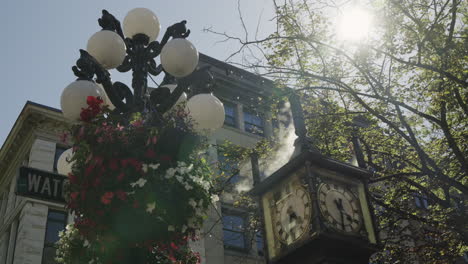 Steam-clock-of-the-Vancouver-Gastown-district,-with-steam-rising,-sun-flares-and-a-feather-flying-through-the-image,-and-a-street-lamp-with-hanging-flowers,-handheld-slow-motion