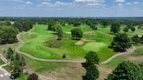 Lush-green-golf-course-with-players-and-pathways,-city-skyline-in-the-distance