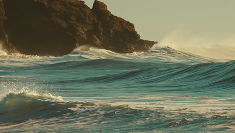 Riesige-Wellen-Rollen-Bei-Sonnenuntergang-über-Die-Küste,-Mit-Schroffen-Klippen-Im-Hintergrund