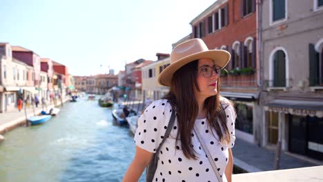 Female-tourist-with-hat-enjoys-sun-on-bridge-over-Canal-in-Murano,-Italy