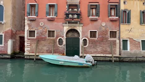 4K-Static-Shot-of-a-Serene-Venetian-Canal:-A-Boat-Drifting-at-the-Entrance-of-a-Magnificent-Historical-Building-in-Venice,-Italy,-Showcasing-Unique-Architecture-and-Rich-Cultural-Heritage