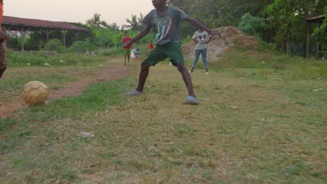 Junger-Mann-Im-Orangefarbenen-Hemd-Dribbelt-Den-Fußball-Durch-Den-Ersten-Jungen,-Der-Zu-Boden-Fiel,-Durch-Die-Beine-Von-Zwei-Anderen-Spielern,-Kumasi,-Ghana