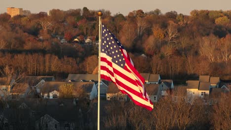 Filmische-Aufnahme-Einer-Amerikanischen-Flagge,-Die-Im-Winter-Während-Des-Sonnenuntergangs-Zur-Goldenen-Stunde-Gegen-Ein-Amerikanisches-Viertel-Inmitten-Kahler-Bäume-Weht