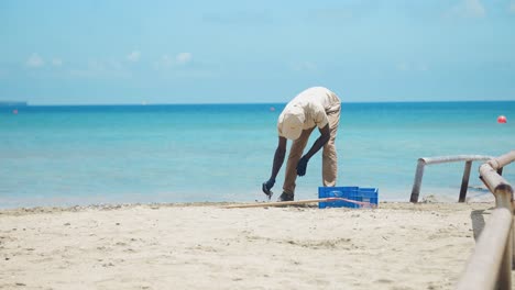 Ein-Schwarzer-Mann-Mit-Baseballkappe-Reinigt-Fleißig-Den-Strand-Mit-Händen-Und-Einem-Rechen,-Sammelt-Müll-Ein-Und-Legt-Ihn-In-Eine-Blaue-Fischkiste