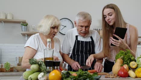 Senior-grandparents-couple-cutting-vegetables-for-salad,-listening-recipe-from-girl-with-tablet