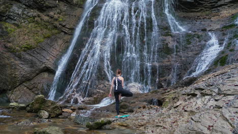 Rear-view-of-a-fit-young-woman-practicing-yoga-in-waterfall-scenery,-wide-shot