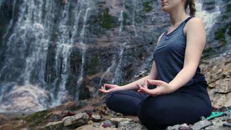 Girl-practicing-flow-yoga-by-a-waterfall,-meditating-in-a-lotus-pose,-handheld