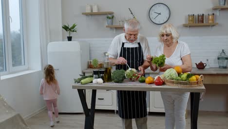 Senior-grandmother-and-grandfather-cooking-salad.-Granddaughter-giving-vegetables-from-refrigerator