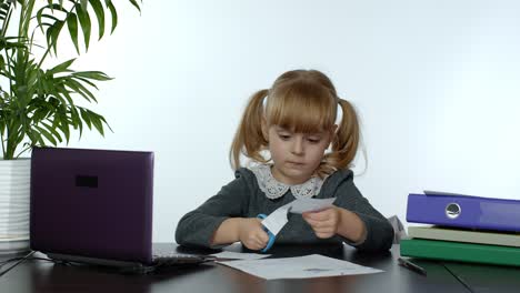 Child-schoolgirl-learns-lessons-at-home-sitting-at-table-cutting-with-scissors-shapes-out-of-paper