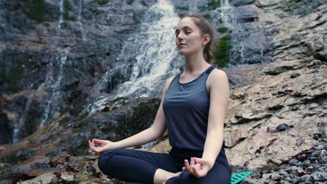 Woman-practicing-yoga,-sitting-in-om-pose-on-the-rock-near-a-waterfall,-handheld