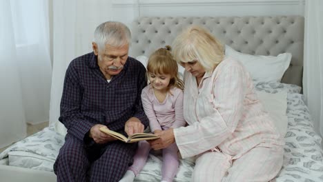 Süßes-Mädchen-Mit-älterer-Großmutter-Und-Großvater-Im-Ruhestand,-Die-Auf-Dem-Bett-Sitzen-Und-Im-Schlafzimmer-Ein-Buch-Lesen