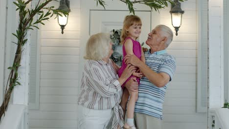 Glückliches-älteres-Großvater--Und-Großmutterpaar,-Das-Enkelin-In-Den-Händen-Auf-Der-Veranda-Zu-Hause-Hält