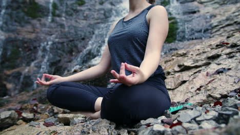 Meditation-in-nature,-a-girl-relaxing-in-a-yoga-pose-near-a-waterfall,-handheld