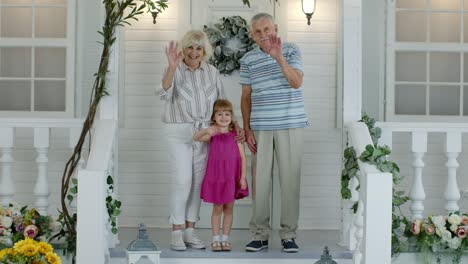 Senior-grandfather-and-grandmother-couple-with-granddaughter-waving-hand,-smiling,-saying-hello