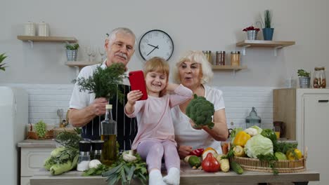 Blogger-girl-taking-photos-on-mobile-phone-with-senior-grandparents-at-kitchen-with-vegetables