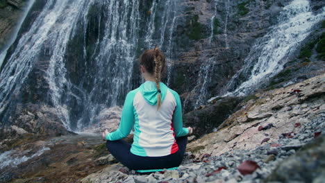 Woman-in-a-lotus-yoga-pose-with-raised-arms-overhead,-by-a-waterfall,-rear-view