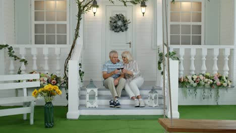 Satisfied-senior-elderly-Caucasian-couple-sitting-and-drinking-wine-in-porch-stairs-at-home