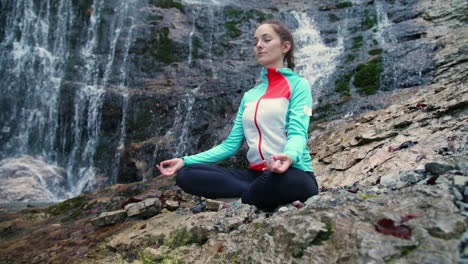Woman-meditating,-sitting-in-lotus-yoga-position-by-a-waterfall,-low-angle-shot