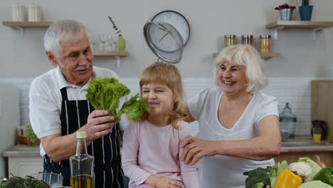 Happy-vegan-senior-couple-dancing-with-granddaughter-child-while-cooking-vegetables-in-kitchen