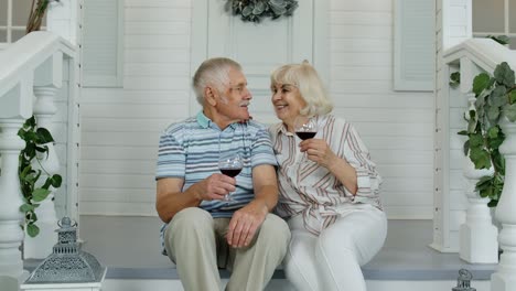 Älteres-Kaukasisches-Paar-Trinkt-Wein-Auf-Der-Veranda-Zu-Hause,-Schaut-In-Die-Kamera-Und-Lächelt